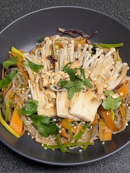 Asiatische Wok-Pfanne mit Konjak-Nudeln, feinen Möhrenstreifen und knusprig gebratenen Enoki-Pilzen, garniert mit frischem Koriander und gehackten Erdnüssen auf einem dunklen Teller.
